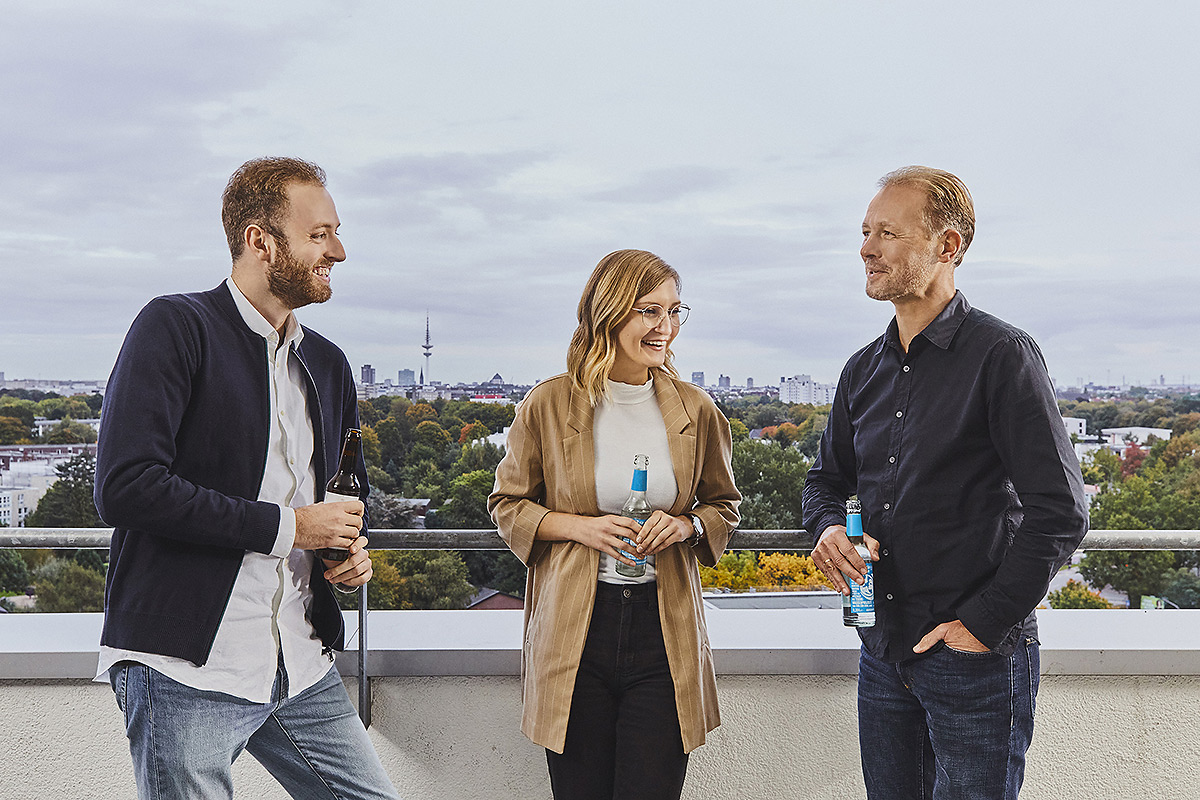 3 Personen stehen auf der Terrasse, trinken Wasser und blicken auf Hamburg.
