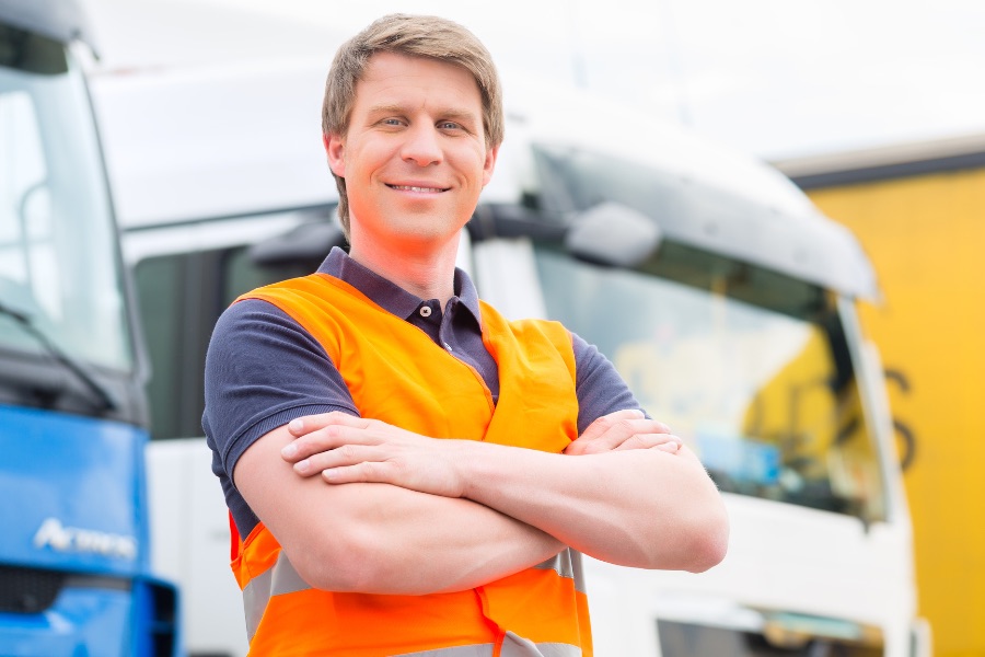 LKW-Fahrer in orangefarbener Sicherheitsweste mit vor der Brust verschränkten Armen vor mehreren Lastwagen.