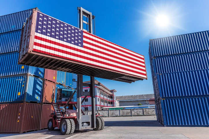 Versand von Deutschland in die USA. Bild zeigt einen Stacker in einem Containerterminal, der einen Container mit der US-Flagge transportiert.